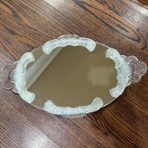 May include: An oval-shaped vanity mirror tray featuring a reflective centre surface surrounded by a clear, pressed glass border. The glass frame displays a decorative scalloped edge with intricate, raised fan-like patterns and textured detailing.