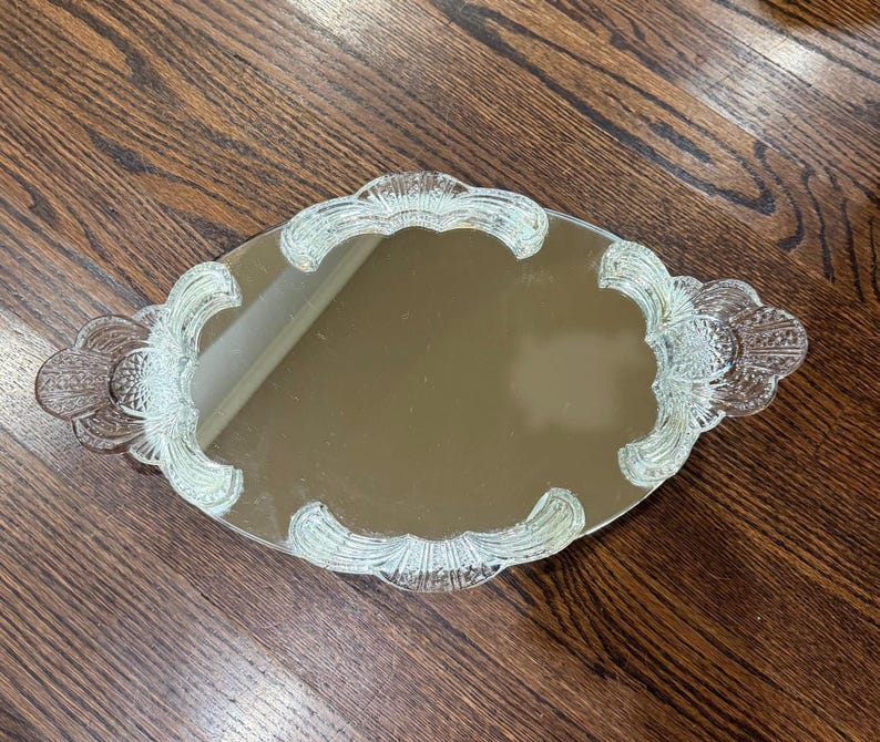 May include: An oval-shaped vanity mirror tray featuring a reflective centre surface surrounded by a clear, pressed glass border. The glass frame displays a decorative scalloped edge with intricate, raised fan-like patterns and textured detailing.