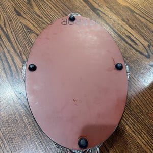 May include: An oval-shaped tray with a smooth, reddish-brown underside featuring four small black rubber feet. The tray has a decorative, textured silver-toned metal border with a scalloped edge and a visible mirror surface on the top side.