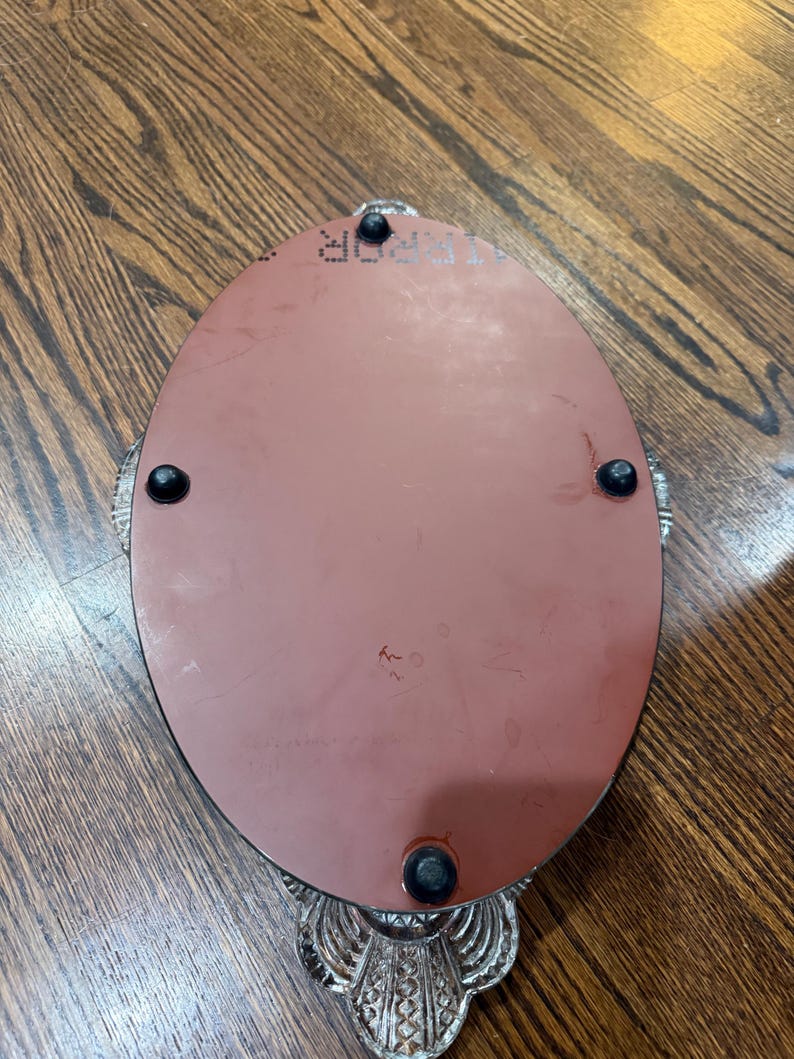 May include: An oval-shaped tray with a smooth, reddish-brown underside featuring four small black rubber feet. The tray has a decorative, textured silver-toned metal border with a scalloped edge and a visible mirror surface on the top side.