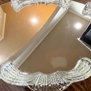 May include: A mirrored vanity tray featuring a reflective glass surface and two clear, pressed glass handles. The handles exhibit a scalloped, fan-like pattern with textured detailing, providing a decorative border on opposite sides of the tray.