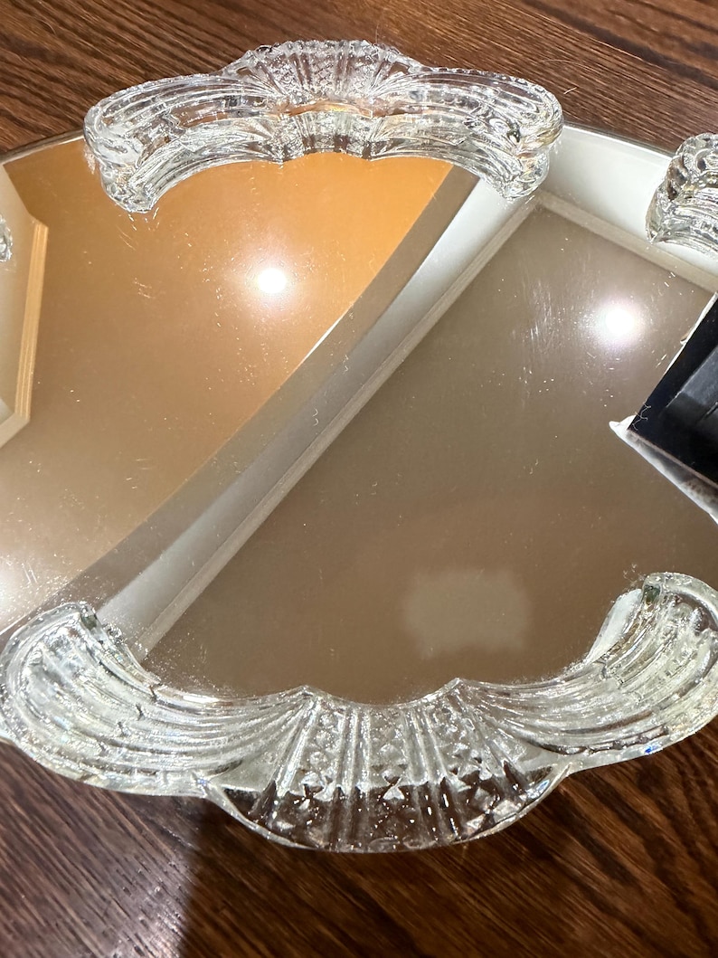 May include: A mirrored vanity tray featuring a reflective glass surface and two clear, pressed glass handles. The handles exhibit a scalloped, fan-like pattern with textured detailing, providing a decorative border on opposite sides of the tray.