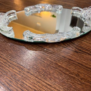 May include: A round vanity mirror tray featuring a reflective glass surface surrounded by a clear, textured glass border. The border consists of four distinct, curved segments with a ribbed, fan-like pattern that frames the central mirror.
