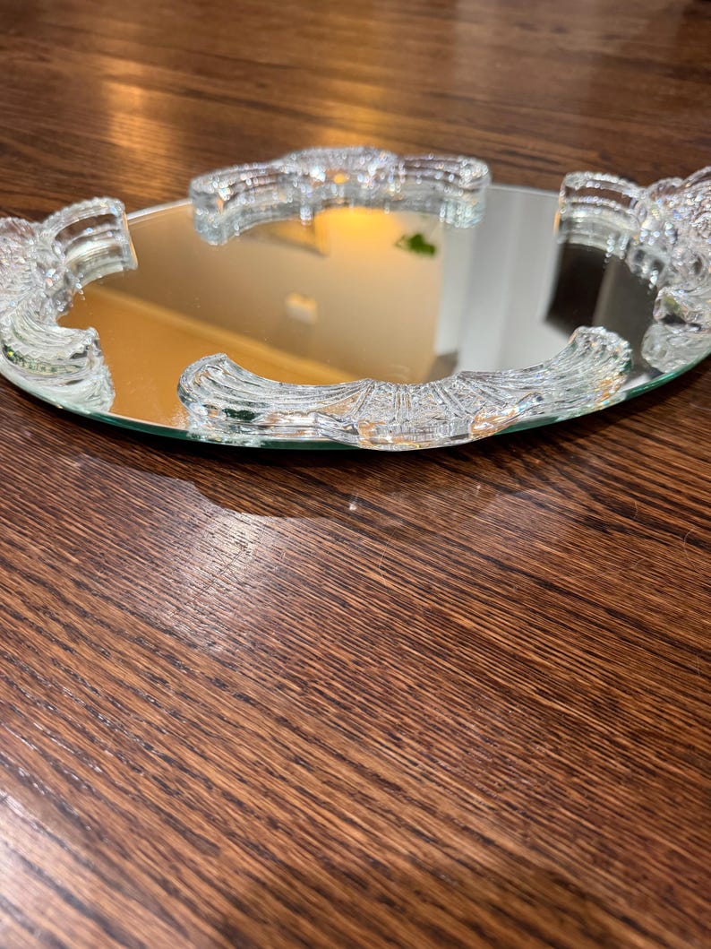 May include: A round vanity mirror tray featuring a reflective glass surface surrounded by a clear, textured glass border. The border consists of four distinct, curved segments with a ribbed, fan-like pattern that frames the central mirror.