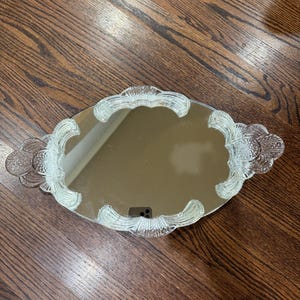 May include: An oval-shaped vanity mirror tray featuring a reflective centre surface surrounded by a clear, textured glass border with decorative scalloped edges and integrated handles on both sides, showcasing a detailed pressed glass pattern.