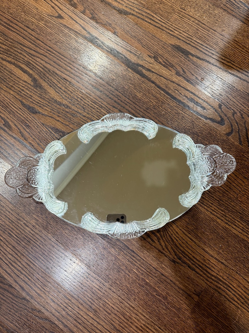 May include: An oval-shaped vanity mirror tray featuring a reflective centre surface surrounded by a clear, textured glass border with decorative scalloped edges and integrated handles on both sides, showcasing a detailed pressed glass pattern.