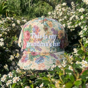 Puede incluir: Gorra de béisbol con estampado floral y el texto bordado "This is my grandma's hat". La gorra presenta un diseño de tapiz colorido con flores rosas, azules y amarillas. El sombrero está rodeado de vegetación y pequeñas flores blancas.