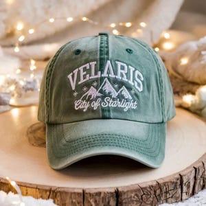 May include: A green baseball cap with the word "VELARIS" embroidered in white above a mountain graphic and the text "City of Starlight". The cap is made of a soft material and has a curved brim.