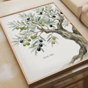 Op de afbeelding: Een ingelijste aquarelprint van een olijfboomtak met groene bladeren en zwarte en groene olijven. Het kunstwerk toont een getextureerde bruine stam en takken tegen een witte achtergrond met de tekst OLIVE TREE in het zwart gedrukt.