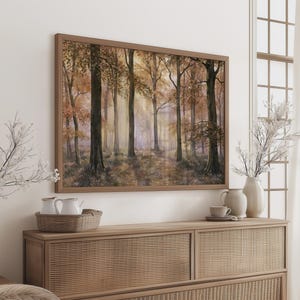 May include: A rectangular framed wall art print depicting a forest scene with tall trees, golden autumn foliage, and soft light filtering through the branches. The artwork is displayed above a wooden cabinet with a woven texture and light wood finish.