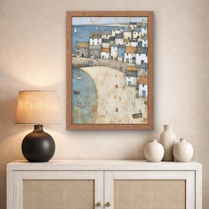 May include: A framed wall art print depicting a coastal village scene with white and blue houses clustered along a sandy beach. The painting features small sailboats on the water, a curved shoreline, and a muted color palette of beige, blue, and grey.