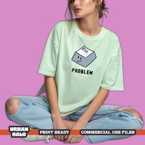 May include: A light green short-sleeve t-shirt featuring a graphic of a gray computer keyboard escape key with a smiling face. Below the key, the word "PROBLEM" is printed in black capital letters. The shirt has a crew neckline and loose fit.