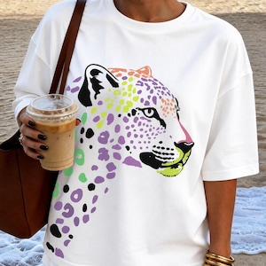 Puede incluir: Una camiseta blanca de manga corta con un gran estampado gráfico de la cabeza y el cuello de un leopardo. El diseño del leopardo está representado de forma estilizada con manchas en tonos morado, verde lima y naranja, y contornos negros.