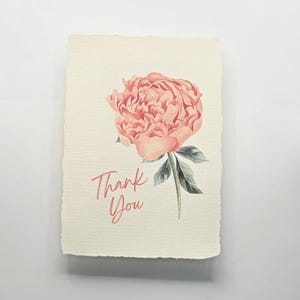 May include: A rectangular greeting card with a deckled edge featuring a watercolor illustration of a large, layered peach-colored peony flower with green leaves. The text Thank You is written in a cursive, coral-colored script on the left side.