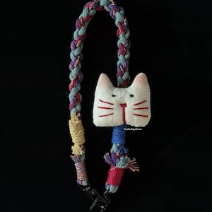 Puede incluir: Un charm de bolso hecho a mano con una cara de gato blanca y detalles rojos. Cuenta con una correa trenzada en tonos azules, morados y rojos, y un cierre negro. El texto "Tactile Bag Charm" es visible.