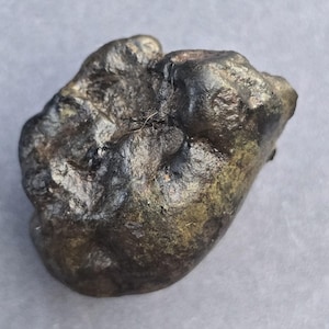 May include: A dark, irregularly shaped stone with a rough, textured surface. The stone has a mix of dark gray, brown, and hints of green hues. The overall shape is organic and natural, suggesting a mineral or geological specimen.