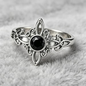 Può includere: Un anello color argento con una pietra preziosa nera rotonda al centro, incastonata in una montatura metallica simmetrica e decorata. La fascia presenta intricati motivi in filigrana che si estendono verso l'esterno a croce.