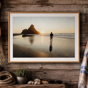 Puede incluir: Fotografía enmarcada de una persona caminando por una playa al atardecer. La imagen presenta una silueta de una persona, una gran formación rocosa y el sol poniéndose sobre el océano. Se ven un sombrero de paja, una red de pesca y una tabla de surf.