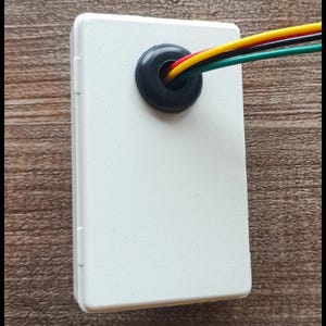 Puede incluir: Una caja rectangular de plástico blanco para componentes electrónicos con un pasacables de goma negro centrado en la parte superior. Cuatro cables aislados de colores amarillo, rojo, negro y verde salen por la abertura del pasacables.