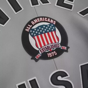 May include: A light gray jacket featuring a circular embroidered patch with a red, white, and blue shield design. The patch text reads ALL AMERICANS ESTABLISHED 1975 N.Y. Large black and white letters are visible at the top and bottom of the garment.