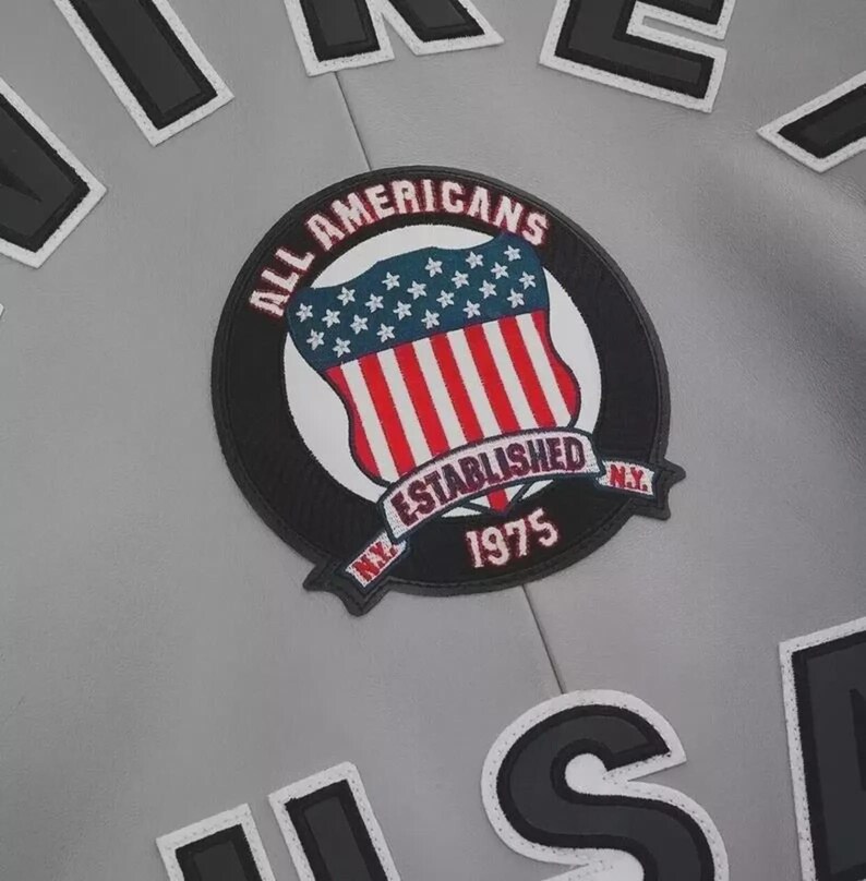 May include: A light gray jacket featuring a circular embroidered patch with a red, white, and blue shield design. The patch text reads ALL AMERICANS ESTABLISHED 1975 N.Y. Large black and white letters are visible at the top and bottom of the garment.