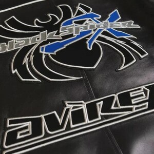 May include: A black leather jacket featuring a large embroidered graphic of a stylized spider with the text Black Spider in silver and blue. Below the spider graphic, the word Avire is embroidered in bold black letters with a white outline.