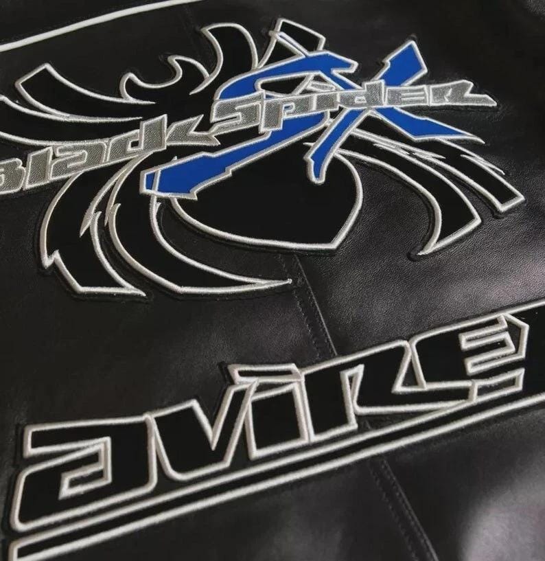 May include: A black leather jacket featuring a large embroidered graphic of a stylized spider with the text Black Spider in silver and blue. Below the spider graphic, the word Avire is embroidered in bold black letters with a white outline.
