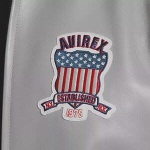 May include: A light gray leather jacket featuring a prominent embroidered patch. The patch displays the text AVIREX in dark blue, a shield with white stars on a blue field and red and white stripes, and a banner reading ESTABLISHED N.Y. 1975.