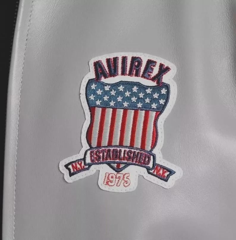 May include: A light gray leather jacket featuring a prominent embroidered patch. The patch displays the text AVIREX in dark blue, a shield with white stars on a blue field and red and white stripes, and a banner reading ESTABLISHED N.Y. 1975.