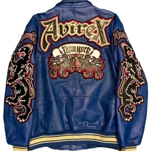 May include: Blue leather jacket with embroidered patches. The back features the word "Avirex" in large letters, a "New York" banner, and two lion emblems. The sleeves have additional lion designs. The jacket has a yellow and white striped trim.
