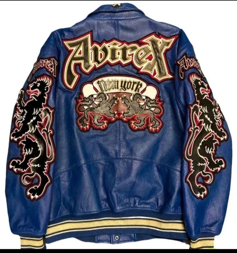 May include: Blue leather jacket with embroidered patches. The back features the word "Avirex" in large letters, a "New York" banner, and two lion emblems. The sleeves have additional lion designs. The jacket has a yellow and white striped trim.