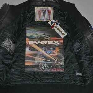 May include: A black leather jacket with a black quilted lining. The interior features a large graphic print of a motorcycle and the text AVIREX. A rectangular brand label with three pennants and a hanging tag with the text AVIREX are attached.