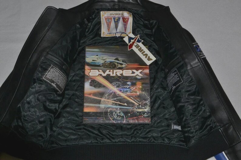 May include: A black leather jacket with a black quilted lining. The interior features a large graphic print of a motorcycle and the text AVIREX. A rectangular brand label with three pennants and a hanging tag with the text AVIREX are attached.