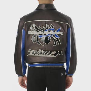 May include: A dark brown leather jacket featuring blue side panels and white piping. The back displays a large, glossy black spider graphic with the blue text "Black Spider" and the white text "Avirex" printed in a bold, stylized font.