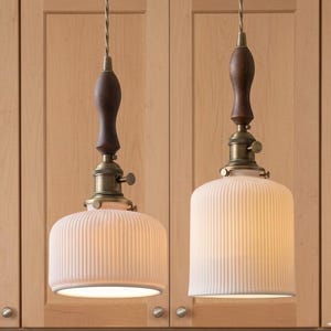 May include: Two pendant lights with a vintage design. Each light has a ribbed, off-white glass shade, a dark brown wooden top, and brass-coloured hardware. The lights hang from a twisted, gold-coloured cord, against a light wood cabinet.