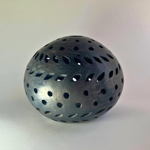 May include: A dark gray, egg-shaped decorative object with numerous leaf-shaped and circular cutouts. The surface has a textured appearance, and the cutouts allow light to pass through, creating interesting patterns. This item is likely a decorative home accent.