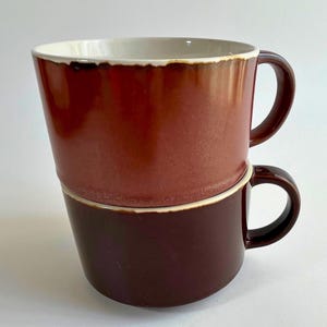 May include: Two stacked ceramic mugs. The top mug is a gradient of reddish-brown, with a white interior and a dark brown handle. The bottom mug is a solid dark brown with a matching handle. Both mugs have a simple, modern design.