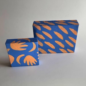 May include: Two rectangular boxes with a blue base color. The smaller box features orange hand-shaped and crescent moon motifs. The larger box is decorated with a repeating pattern of diagonal orange brushstroke marks across the blue surface.