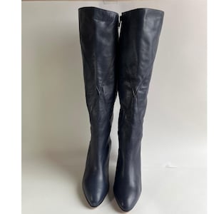May include: A pair of knee-high boots in a dark navy blue leather. These boots feature a pointed toe, a slim heel, and a distinct V-shaped seam detail running down the front of the shaft. The leather has a smooth finish with subtle natural creasing.