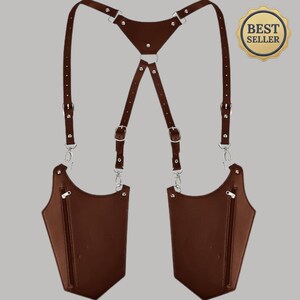 May include: A brown leather shoulder holster bag featuring two symmetrical pockets with vertical silver-tone zippers. The harness system includes adjustable straps with metal buckles and a central triangular back piece connected by silver-tone hardware.