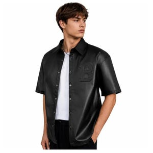 Pode incluir: Uma camisa preta de manga curta com botões, feita de um material liso semelhante a couro. A camisa apresenta uma letra B proeminente em relevo no lado esquerdo do peito e é usada aberta sobre uma t-shirt branca de cor sólida.