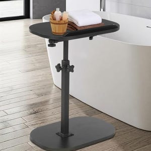 May include: A black adjustable bath tray with a rectangular top and an oval base. The tray holds a small wooden bucket, bottles, and folded white and brown towels. The stand is positioned next to a white bathtub.