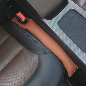 May include: A brown car seat gap filler is shown, designed to prevent items from falling into the space between the car seat and the center console. The filler is a long, slender shape, made of a smooth material, and is placed next to a seat belt buckle.