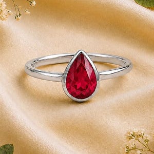 May include: A silver-toned ring featuring a single pear-shaped, faceted red gemstone set in a smooth bezel setting. The band is polished and slender, showcasing a minimalist design that highlights the vibrant crimson colour of the central stone.