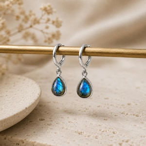Puede incluir: Un par de pendientes de aro de plata con piedras preciosas de labradorita en forma de lágrima. Cada pendiente consta de un aro de plata pulida con cierre de bisagra y una piedra de labradorita facetada que muestra iridiscencia azul.