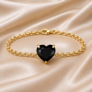 May include: A gold chain bracelet featuring a single, faceted, heart-shaped black gemstone set in the centre. The bracelet is constructed with circular links and includes a gold lobster claw clasp at the top for secure fastening.
