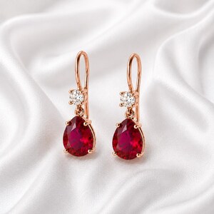 Puede incluir: Un par de pendientes colgantes de oro rosa con una piedra preciosa roja en forma de pera suspendida de una pequeña piedra redonda transparente. Los pendientes tienen un cierre de gancho curvado y un acabado de metal pulido brillante.