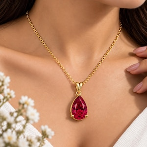 May include: A gold chain necklace featuring a pear-shaped, faceted deep red gemstone pendant. The vibrant red stone is set in a polished gold metal frame with a simple bail, suspended from a delicate gold link chain that rests against the collarbone.
