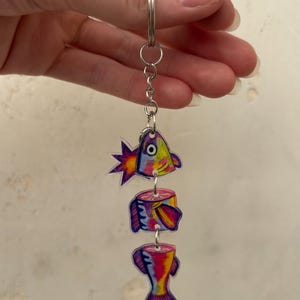 May include: A colorful fish-shaped keychain with three connected pieces. The fish is segmented into head, body, and tail sections, each with vibrant hues of pink, yellow, blue, and purple. The keychain is attached to a silver metal ring.