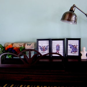 May include: Three framed prints of bats with musical notes in the background. The prints are on a wooden surface with a lamp and a decorative box with flowers.